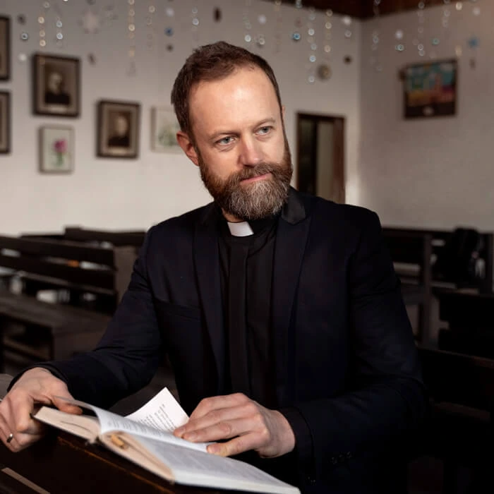 A young woman wearing glasses and a beige sweater reads a book. Blurred people are in the background